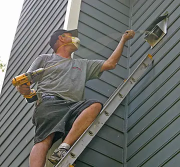 CleanFlow Pros technician on a ladder servicing an exterior dryer vent hood with a cordless drill
